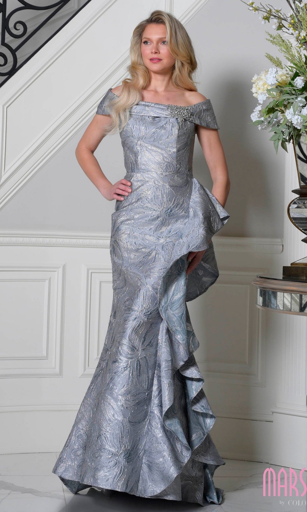 A woman stands indoors in the Long Metallic Brocade Formal Dress: Marsoni MV1381, featuring an off-the-shoulder silver textured design with a ruffled side—an elegant pick for mothers of the bride or any special evening occasion.